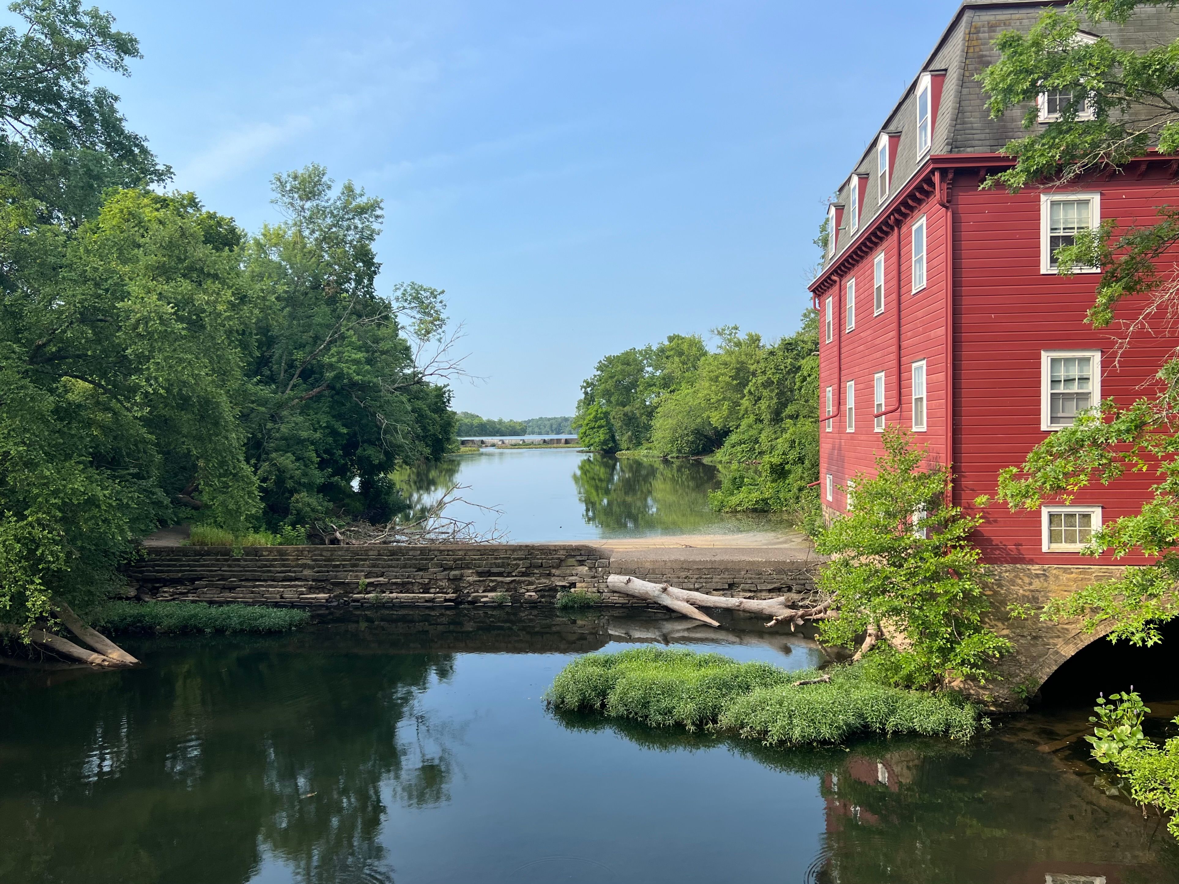 Old red mill house sits on a lake with a wide open vista. Blue skies and green trees.